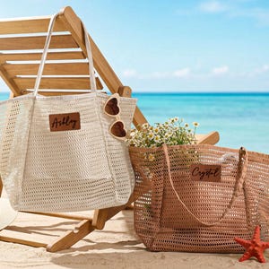 Puede incluir: Dos bolsas de playa de malla, una crema y otra marrón, están en una playa de arena cerca de una silla de playa de madera. La bolsa crema tiene el nombre "Ashley" en un parche de cuero. La bolsa marrón tiene el nombre "Crystal".