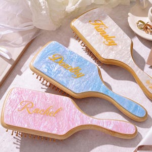 May include: Three rectangular hairbrushes with wooden handles and pearlescent acrylic backs in pink, blue, and white. Each brush features a custom name in gold cursive script: Rachel on the pink brush, Dorothy on the blue, and Kinsley on the white.