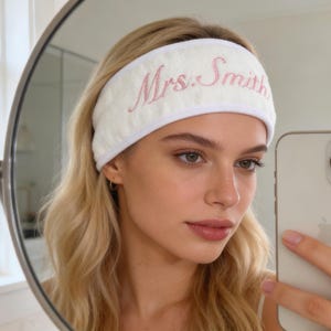 Op de afbeelding: Witte badstof hoofdband geborduurd met de naam "Mrs. Smith" in roze script. De hoofdband wordt gedragen door een persoon met blond haar, zichtbaar in een spiegel selfie. De hoofdband heeft een textuurpatroon en een witte rand.