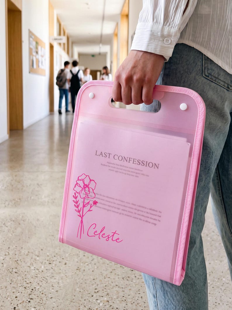 K&ouml;nnte beinhalten: Eine halbtransparente rosa Dokumentenmappe aus Kunststoff mit integriertem Griff und zwei wei&szlig;en Druckkn&ouml;pfen. Auf der Vorderseite befindet sich eine florale Strichzeichnung, der Text "LAST CONFESSION" in Serifenschrift und der Name "Celeste" in Schreibschrift.