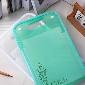 K&ouml;nnte beinhalten: Eine durchscheinende mintgr&uuml;ne Dokumentenmappe aus Kunststoff mit einem Tragegriff und zwei wei&szlig;en Druckkn&ouml;pfen. Die Mappe ist mit einer dunkelgr&uuml;nen Strichzeichnung eines Blumenzweigs und dem Namen Natalie in Schreibschrift unten bedruckt.
