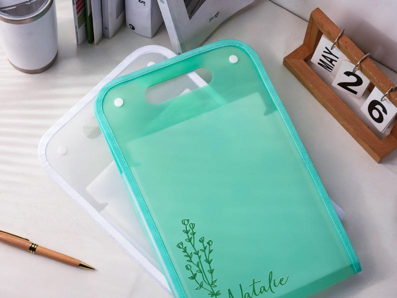 K&ouml;nnte beinhalten: Eine durchscheinende mintgr&uuml;ne Dokumentenmappe aus Kunststoff mit einem Tragegriff und zwei wei&szlig;en Druckkn&ouml;pfen. Die Mappe ist mit einer dunkelgr&uuml;nen Strichzeichnung eines Blumenzweigs und dem Namen Natalie in Schreibschrift unten bedruckt.