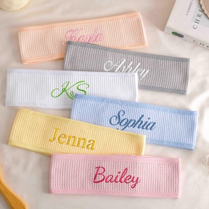 May include: A collection of personalized headbands in various pastel colors, including peach, gray, white, light blue, yellow, and pink. Each headband features an embroidered name in a complementary color, such as "Paula," "Ashley," and "Jenna."