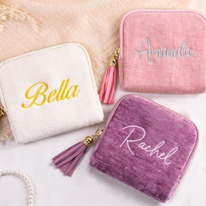 May include: Four personalised square zipped pouches in white, pink, and purple, each embroidered with a name in a contrasting colour. The pouches have gold-toned zips and matching tassels. A pearl necklace and gold rings are also visible.
