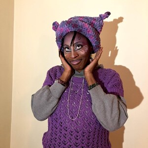 May include: A purple crochet hat with ear-like protrusions, paired with a matching purple short-sleeved sweater over a gray sweatshirt. The sweater has a detailed openwork design. The person is wearing face paint.