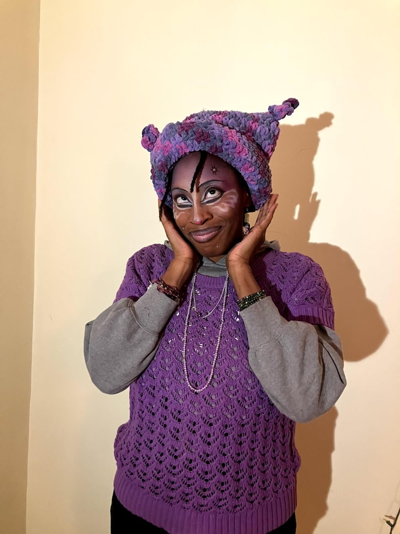 May include: A purple crochet hat with ear-like protrusions, paired with a matching purple short-sleeved sweater over a gray sweatshirt. The sweater has a detailed openwork design. The person is wearing face paint.