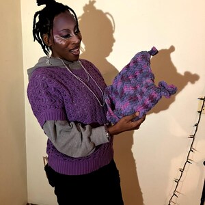 May include: A handmade, textured hat in shades of purple and pink is held up. The hat appears to be crocheted or knitted. The person holding the hat is wearing a purple sweater over a gray long-sleeved shirt and black pants.