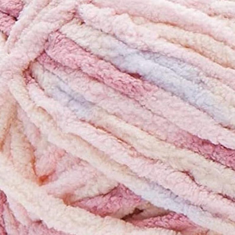 May include: Close-up of a skein of soft, plush yarn in shades of pink, cream, and lavender. The yarn has a thick, textured appearance, ideal for knitting or crocheting projects. The colors blend together, creating a gradient effect.
