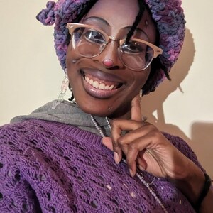 May include: A person wearing a purple crochet sweater, a matching purple and pink beret, and gold-rimmed glasses. The sweater has a detailed openwork design. The person is also wearing a silver necklace and earrings.