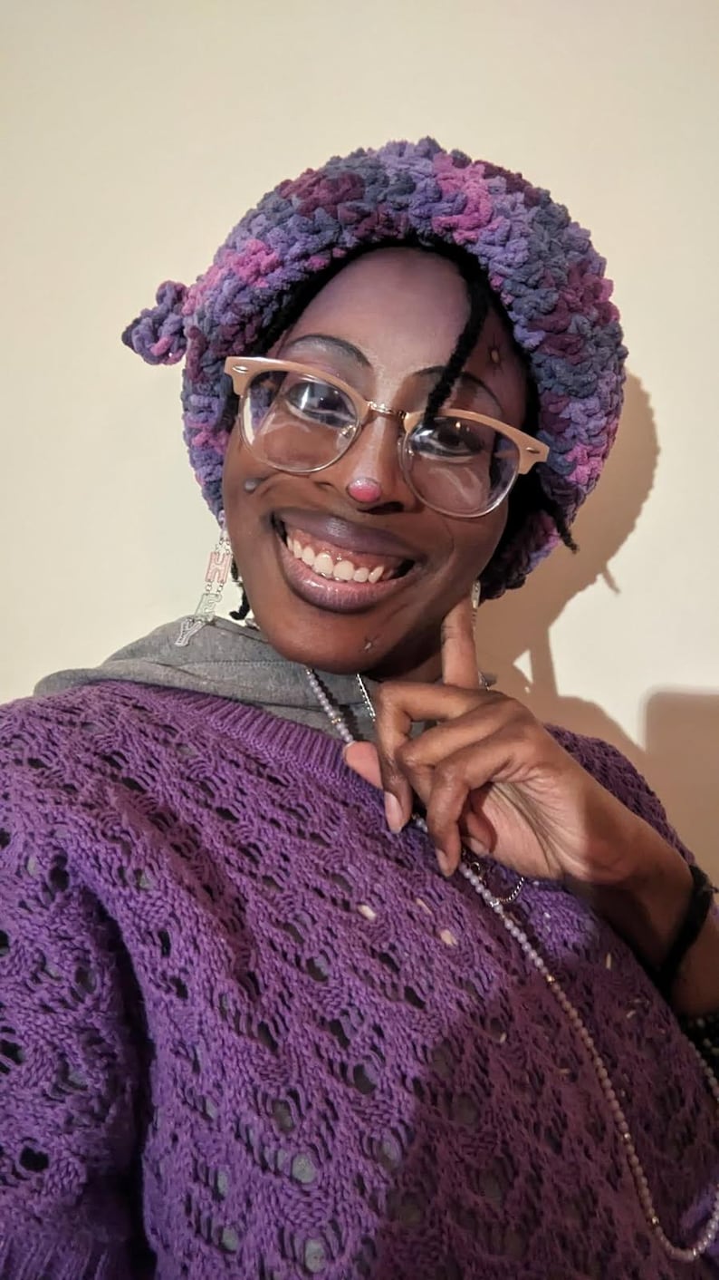 May include: A person wearing a purple crochet sweater, a matching purple and pink beret, and gold-rimmed glasses. The sweater has a detailed openwork design. The person is also wearing a silver necklace and earrings.