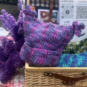 May include: A selection of handmade crochet items, including a purple and pink hat with decorative knots, and a matching scarf. Other items include a purple, plush, decorative item and a blue and green scarf. The items are displayed in a wicker basket.