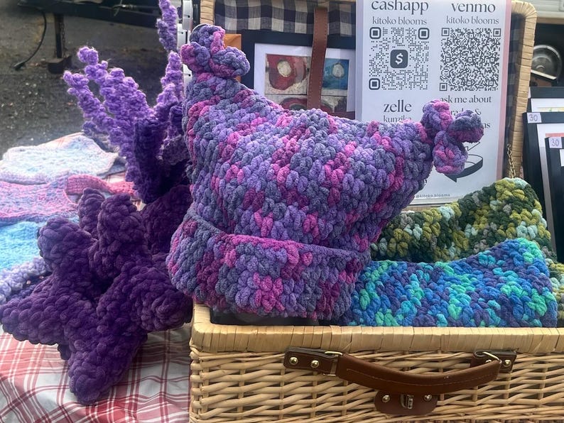May include: A display of handmade crochet items, including a purple and pink hat with decorative knots, and a matching scarf. Other items include a purple, plush, decorative item and a blue and green scarf. The items are displayed in a wicker basket.
