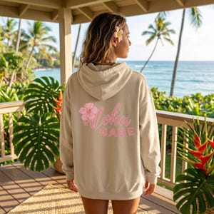 May include: A tan hoodie with a drawstring hood and a pink floral design with the words "Aloha Babe" printed on the back. The hoodie is made of a soft material and is perfect for a casual, beachy look.