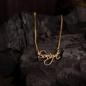 Könnte beinhalten: Eine goldfarbene Halskette mit dem Namen "Soniya" in Schreibschrift. Die Kette wird vor einem dunklen, strukturierten Hintergrund präsentiert, der das zarte Design und den warmen Farbton des Schmucks hervorhebt.
