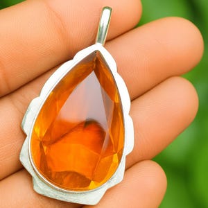 Könnte beinhalten: Ein tropfenförmiger Anhänger mit einem großen, facettierten, orangefarbenen Edelstein. Der Stein ist in einen silberfarbenen Metallrahmen mit dekorativem Rand und einer kleinen Schlaufe für eine Kette gefasst. Der Hintergrund ist ein verschwommenes Grün.