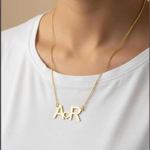 Könnte beinhalten: Eine goldfarbene Halskette mit einer zarten Kette und einem Anhänger, der aus den Großbuchstaben A und R besteht, die durch ein kleines, mittiges Herz-Symbol getrennt sind. Der Anhänger hängt an einer dünnen, polierten Metallkette am Hals.