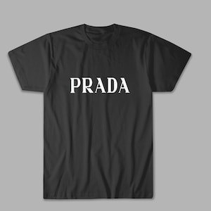 Könnte beinhalten: Schwarzes T-Shirt mit dem weißen Wort "PRADA" auf der Brust. Das Shirt hat kurze Ärmel und einen Rundhalsausschnitt. Der Hintergrund ist einfarbig hellgrau.