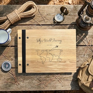 May include: A wooden photo album with the text "Tilly's World Journey" and a world map outline. The album is surrounded by a compass, a lantern, a mug, a backpack, and rope on a wooden surface.