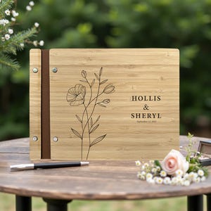 May include: A wooden photo album featuring a floral design and the names "HOLLIS & SHERYL" with the date "September 13, 2025" engraved. The album has a brown leather spine. It is open on a wooden table.