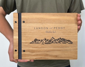 Peut inclure: Un livre d'aventure en bois avec les noms "LANDON and PEGGY" et les mots "Adventure book" gravés sur la couverture. Un motif de chaîne de montagnes est également gravé. Le livre a une tranche bleu foncé et des rivets noirs.