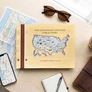 Pode incluir: Um diário de viagem de madeira com o texto "OUR ADVENTURES TOGETHER Sally & Sheldon" e um mapa dos EUA. O diário está aberto numa superfície de madeira clara com um mapa, óculos de sol, um telemóvel, um caderno e uma chávena de café.