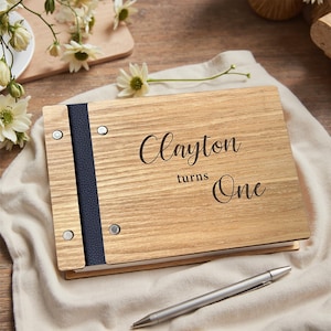 May include: A wooden photo album engraved with "Clayton turns One". The album features a navy blue spine and silver rivets. A silver pen lies next to the album. The album is made of light-colored wood.