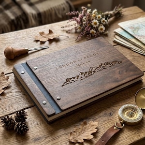 Peut inclure: Un album photo rectangulaire en bois avec une finition brun foncé, présentant une chaîne de montagnes gravée au laser et le texte "LANDON and PEGGY Adventure book" sur la couverture. L'album est fixé par des rivets en métal argenté.