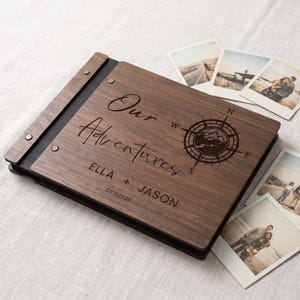 Peut inclure: Un album photo rectangulaire en bois avec une finition grainée brun foncé et une reliure en cuir noir fixée par des rivets métalliques. La couverture est gravée avec le texte Our Adventures, une boussole, les noms ELLA + JASON et EST. 2020.
