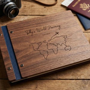 Może przedstawiać: Drewniany album na zdjęcia z napisem "Sibby's World Journey" i wzorem mapy świata. Album ma niebieski skórzany grzbiet i leży na drewnianej powierzchni. Widoczny jest również zabytkowy aparat fotograficzny, paszport i kompas.