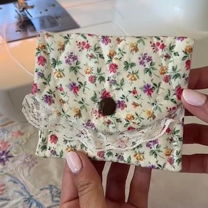 May include: A small quilted fabric pouch featuring a cream background with a colorful floral pattern of pink, purple, and yellow flowers. The pouch has a scalloped flap edge trimmed with white lace and a single dark brown circular button closure.