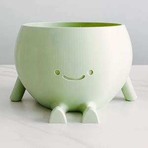 May include: A light green, rounded planter with a smiling face design. The planter has small, rounded legs and a cheerful expression. The interior is a darker shade of green. A decorative item for plants.