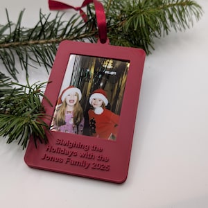 May include: A burgundy Christmas photo frame with a picture of two children in Santa hats. The frame has the text "Sleighing the Holidays with the Jones Family 2025" and is suspended by a red ribbon.