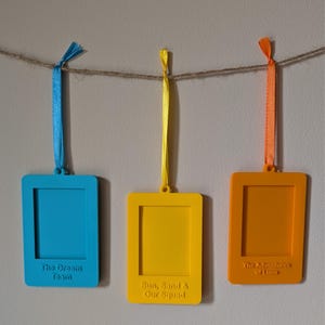 May include: Three brightly coloured picture frame ornaments suspended from a twine string. The frames are blue, yellow, and orange, each with a matching ribbon. The blue frame reads "The Dream Team", the yellow frame reads "Sun, Sand & Our Squad", and the orange frame reads "The Adventures of Luna".
