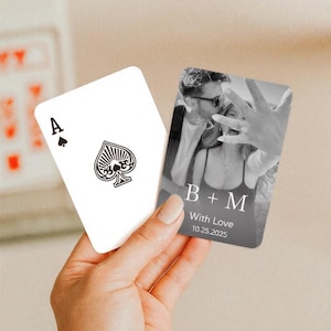 May include: A hand holds two playing cards. One card is the Ace of Spades, the other features a black and white photo with the text "B + M With Love 10.25.2025". The cards are likely for a wedding or engagement.