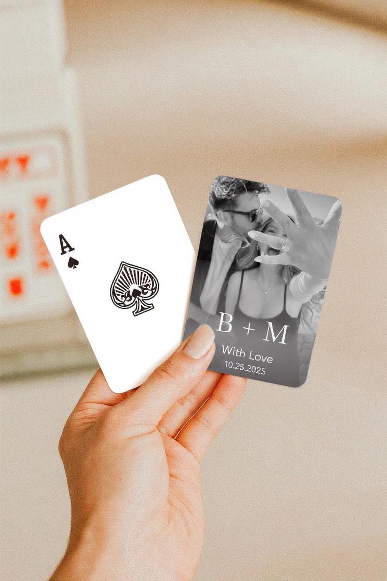 May include: A hand holds two playing cards. One card is the Ace of Spades, the other features a black and white photo with the text "B + M With Love 10.25.2025". The cards are likely for a wedding or engagement.