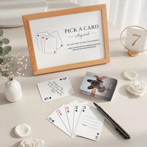 May include: A wooden framed sign with the text "PICK A CARD - Any card" sits next to a deck of cards and a pen. The cards are white with black and red suit symbols. A small vase with white flowers is also present.