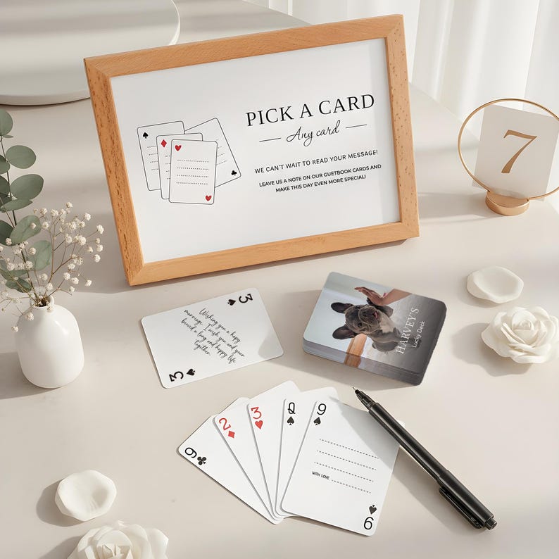 May include: A wooden framed sign with the text "PICK A CARD - Any card" sits next to a deck of cards and a pen. The cards are white with black and red suit symbols. A small vase with white flowers is also present.
