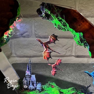 Puede incluir: Una obra de arte de resina de tem&aacute;tica fant&aacute;stica con dos dragones rojos y naranjas volando sobre un castillo en miniatura y un paisaje verde. La obra de arte incluye un borde oscuro con aspecto de madera con acentos verdes brillantes. El texto "Resin Boys" es visible.
