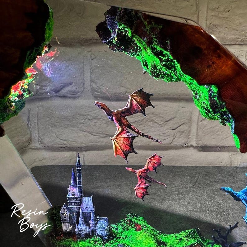 Puede incluir: Una obra de arte de resina de tem&aacute;tica fant&aacute;stica con dos dragones rojos y naranjas volando sobre un castillo en miniatura y un paisaje verde. La obra de arte incluye un borde oscuro con aspecto de madera con acentos verdes brillantes. El texto "Resin Boys" es visible.