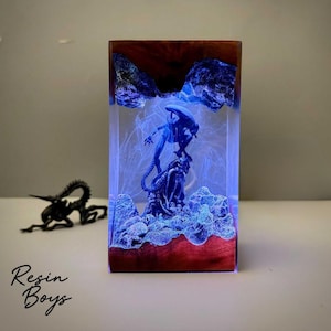May include: A rectangular resin sculpture featuring an Alien figure atop a rock formation, encased in clear resin with a blue glow. The base and top are a dark wood. A small black Alien figure is in the background. The text "Resin Boys" is at the bottom.