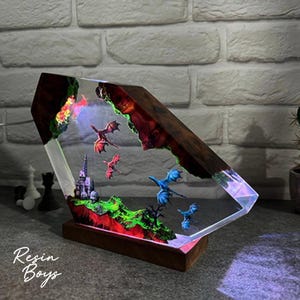 Puede incluir: Una pieza de arte de resina &uacute;nica que presenta una escena de fantas&iacute;a con dragones, un castillo y un paisaje. La obra de arte est&aacute; encerrada en un bloque transparente en &aacute;ngulo con un marco y una base de madera. La pieza est&aacute; iluminada con luces LED de colores.