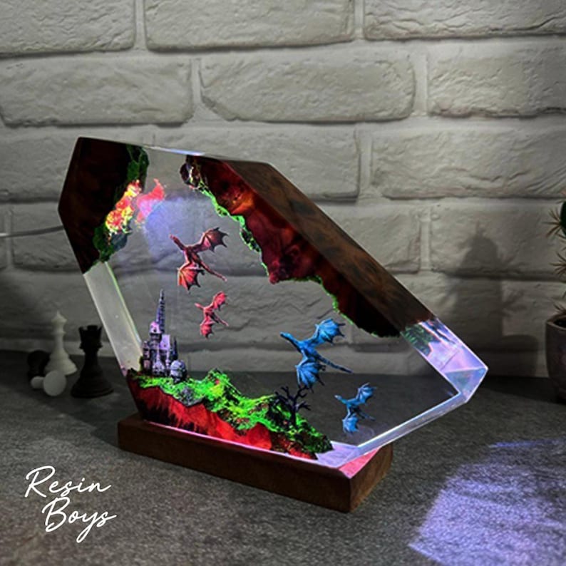 Puede incluir: Una pieza de arte de resina &uacute;nica que presenta una escena de fantas&iacute;a con dragones, un castillo y un paisaje. La obra de arte est&aacute; encerrada en un bloque transparente en &aacute;ngulo con un marco y una base de madera. La pieza est&aacute; iluminada con luces LED de colores.