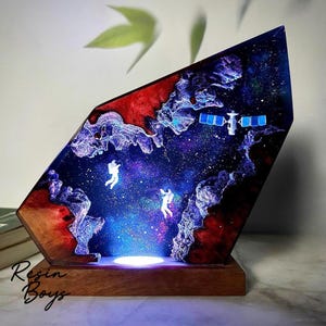 Puede incluir: Una pieza de arte de resina única que presenta una escena espacial con astronautas, un satélite y un efecto de nebulosa. La obra de arte está sobre una base de madera y tiene una fuente de luz en la parte inferior. El texto "Resin Boys" es visible.