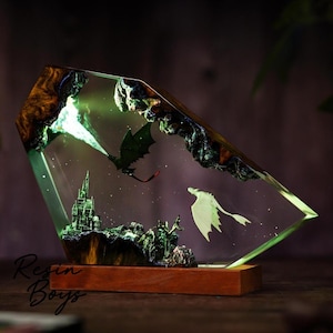 Puede incluir: Escultura de resina fantástica que representa un dragón que lanza fuego verde hacia un castillo. Un dragón blanco vuela cerca. La obra de arte está encerrada en una forma geométrica transparente, montada sobre una base de madera. El texto "Resin Boys" es visible.