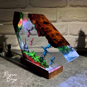 Puede incluir: Una pieza de arte de resina &uacute;nica que presenta una escena de fantas&iacute;a con dragones, un castillo y paisajes coloridos. La obra de arte est&aacute; encapsulada en resina transparente, montada sobre una base de madera, con el texto "Resin Boys".