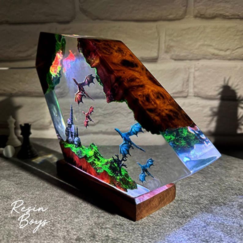 Puede incluir: Una pieza de arte de resina &uacute;nica que presenta una escena de fantas&iacute;a con dragones, un castillo y paisajes coloridos. La obra de arte est&aacute; encapsulada en resina transparente, montada sobre una base de madera, con el texto "Resin Boys".
