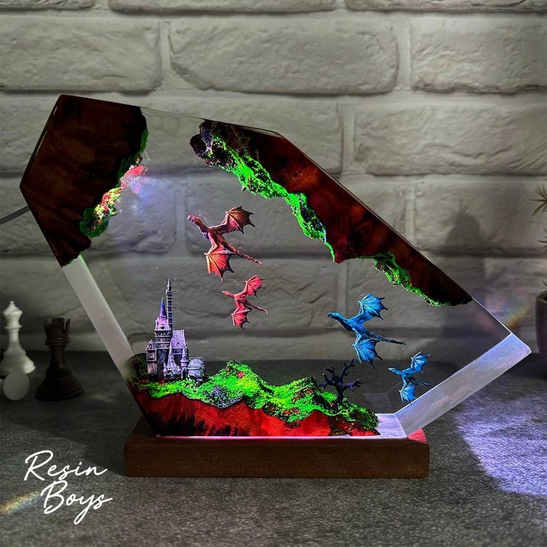 Puede incluir: L&aacute;mpara de resina de fantas&iacute;a con un castillo, monta&ntilde;as y dragones voladores. La l&aacute;mpara tiene una base de madera y un cuerpo de resina transparente y en &aacute;ngulo con detalles coloridos incrustados.