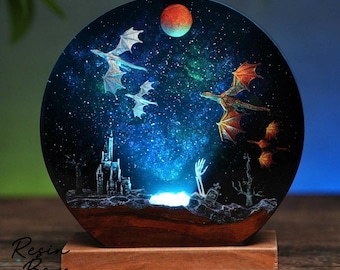 Op de afbeelding: Een ronde hars nachtlamp met een fantasie scène met draken, een kasteel en een begraafplaats onder een sterrenhemel. De draken hebben oranje en witte vleugels. De basis is van hout en het licht gloeit blauw. De tekst "Resin Boys" is zichtbaar.