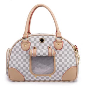 May include: A pet carrier bag featuring a white and gray checkered pattern with tan leather-like trim, gold-tone hardware, a mesh ventilation window with a flap, and two top handles. The bag includes a small circular opening and side accents.