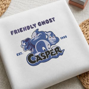 May include: A white sweatshirt featuring an embroidered graphic of three blue Casper ghosts. The text reads "FRIENDLY GHOST" at the top and "EST. 1955" on the sides. The word "CASPER" is prominently displayed in large black letters at the bottom.